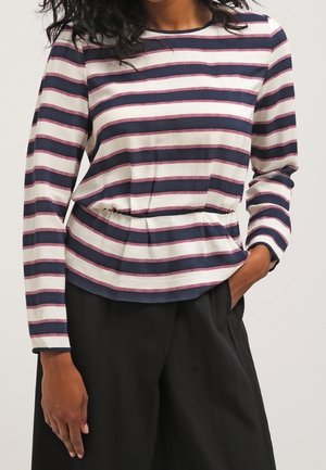 Vrouw draagt een shirt met lange mouwen in marineblauw en wit gestreept met dunne rode details en zwarte wijde broek, hand in de zak.