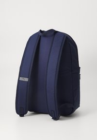 Marineblauwe stoffen rugzak met dikke, gewatteerde schouderbanden en een handvat bovenaan. Voorzien van een grijze logopatch aan de voorkant. Zachte textuur.