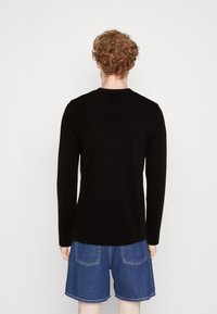 Mann mit lockigem roten Haar, der ein schwarzes Langarmshirt und eine blaue Jeansshorts trägt, steht mit dem Rücken zu einem schlichten weißen Hintergrund.