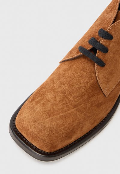 Zapato de ante marrón con puntera cuadrada, cordones negros y costuras en contraste a lo largo del borde. La textura es suave y ligeramente cepillada.