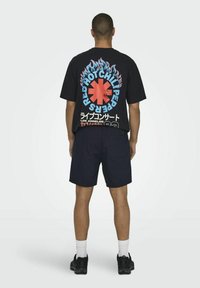 T-shirt noir à manches courtes avec un graphisme coloré représentant des flammes et un texte au dos, associé à un short bleu marine et des baskets noires.
