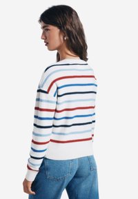 Gestreifter Pullover in Weiß mit blauen, roten und marineblauen Streifen. Stricktextur, Rundhalsausschnitt sowie gerippte Bündchen und Saum. Getragen mit blauen Jeans.