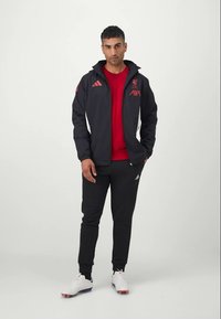 Veste de sport noire avec des accents rouges et le logo de Liverpool, associée à un t-shirt rouge et un pantalon de jogging noir. Chaussures de sport blanches avec des détails colorés.