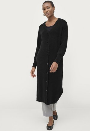 Schwarzer langer Cardigan aus strukturiertem Strick mit V-Ausschnitt, langen Ärmeln und Knopfverschluss. Getragen über grauen Hosen und schwarzen Schuhen.