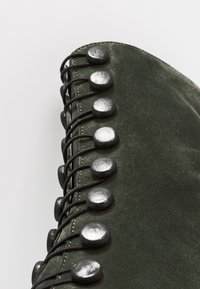 Zelená semišová bota s metalickými stříbrnými knoflíky a černými šňůrkami. Detailní záběr, který zdůrazňuje texturální detaily a švy.