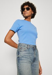 Lys blå tettsittende t-skjorte med korte ermer, kombinert med høyt liv, falmede blå jeans, med en liten logo på brystlommen.