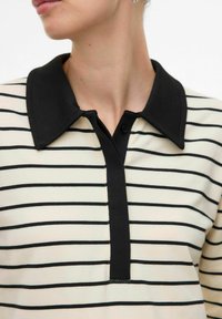 Polo crema con strisce nere, dotato di colletto nero, patta con tre bottoni e morbida texture per il massimo comfort.