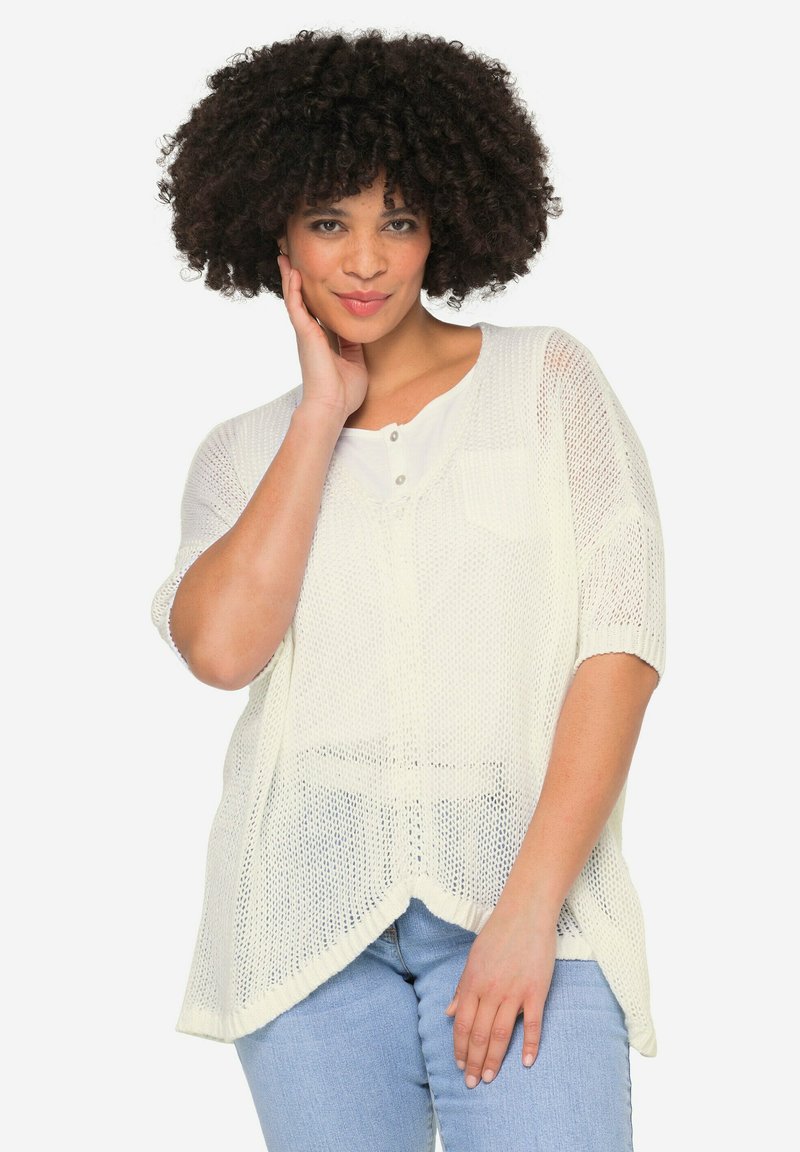 Hvid, let strikket sweater med korte ærmer, rund halsudskæring, sideslids og en frontlomme, båret over blå jeans.