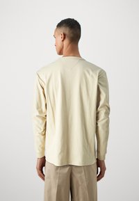 Beige hosszú ujjú póló, puha pamutból készült, kerek nyakkivágással és diszkrét vállvarrással. Világos színű nadrággal párosítva.