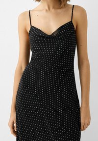 Czarna sukienka bez rękawów z dekoltem w kształcie kapturem, zdobiona białymi kropkami o różnych rozmiarach, wykonana z gładkiego materiału o lekko błyszczącej teksturze.
