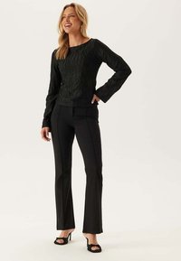 Donna sorridente, che indossa un top nero a maniche lunghe con texture, pantaloni neri attillati e sandali neri con tacco alto e punta aperta, in piedi con una mano sulla vita.