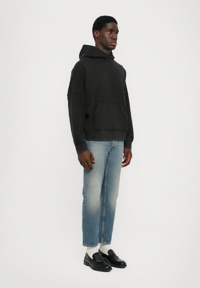 Schwarzer Kapuzenpullover mit einer Fronttasche, kombiniert mit hellblauen Jeans und schwarzen Slippern. Der Stoff wirkt weich und leicht strukturiert.