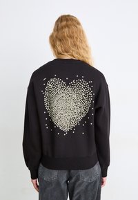 Felpa nera con un grande design a forma di cuore realizzato con piccoli punti bianchi sul retro. Include polsini e orlo a coste.