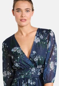 Blumenkleid mit tiefem V-Ausschnitt, Puffärmeln und tailliertem Bund. Aus blauem Stoff mit aufwendigen weißen und grünen Blumenmustern gestaltet.