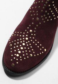 Gros plan sur le bout d'une chaussure en daim bordeaux avec des clous ronds dorés disposés en motif décoratif et un bord de semelle noir.