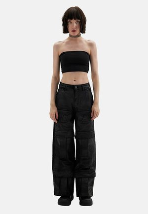 Femme aux cheveux courts et foncés portant un top tube noir, un pantalon cargo noir ample, des chaussures noires et un collier ras-du-cou, debout devant un fond uni.