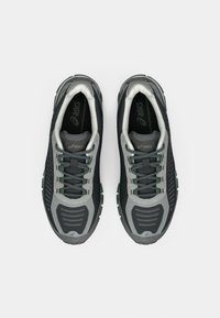 Fekete és szürke sportcipők hálós és szintetikus felsőrésszel, mintás díszítéssel és zöld részletekkel. Kerek fűzővel és gumitalppal rendelkezik.