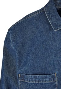Épaule et col d'une veste en denim bleu avec des coutures visibles et une poche avant sur fond blanc.