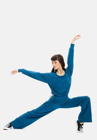 Completo sportivo teal con top morbido e pantaloni a gamba larga. Modella che indossa sneakers nere, in una posa dinamica su uno sfondo neutro.