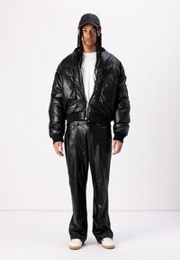 Jachetă bomber oversized din piele sintetică neagră, cu manșete ribbate, asortată cu pantaloni ajustați. Un tricou alb și teniși bej completează ținuta.