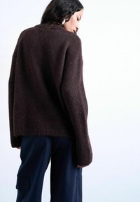 Femme aux longs cheveux foncés portant un pull ample en laine marron foncé et un pantalon cargo bleu marine, regardant vers le bas sur un fond blanc.