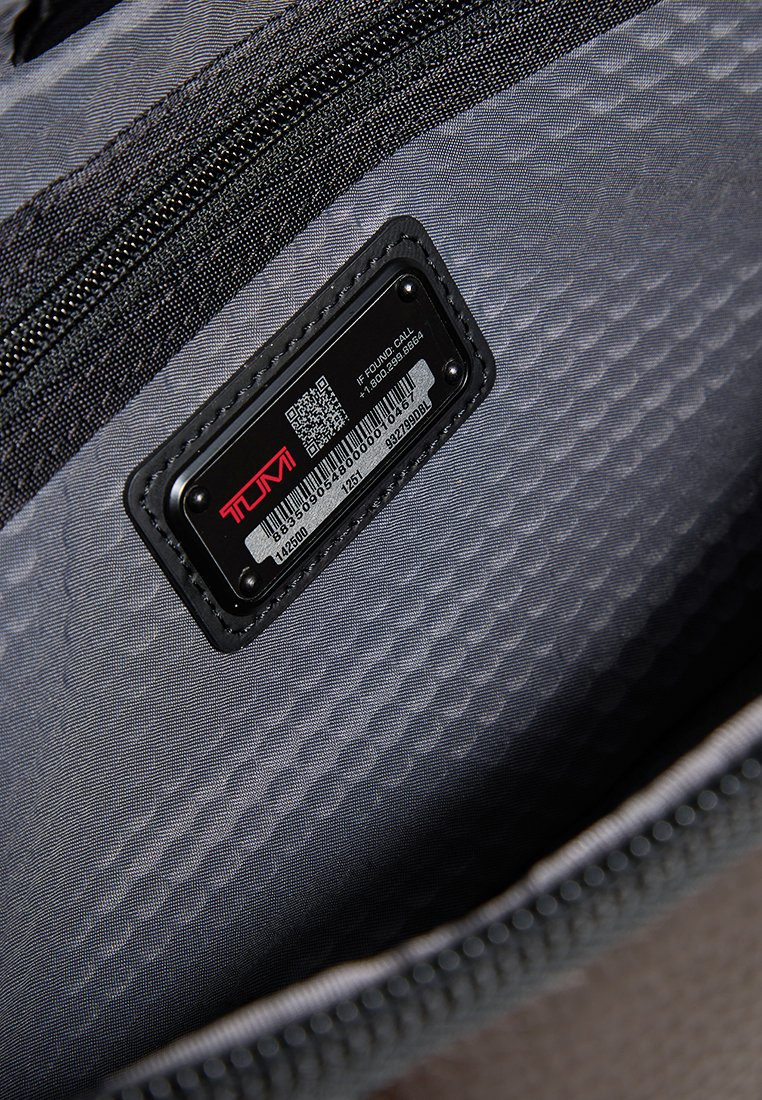 Innenraum einer grauen Stofftasche mit strukturiertem Muster, ausgestattet mit einem schwarzen Identifikationsschild und einem Reißverschluss.