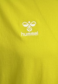 Geel katoenen T-shirt met een wit geborduurd logo met abstract ontwerp. De stof heeft een gladde, gelijkmatige textuur.