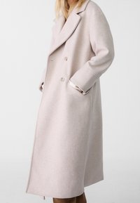 Donna che indossa un lungo cappotto di lana beige chiaro con ampi rever e bottoni doppi, mani nelle tasche, su uno sfondo neutro.