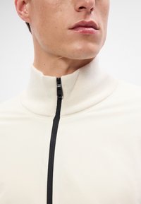 Beige magasnyakú, cipzáros felső sima anyagból; elöl fekete cipzárral és bordázott akcentusokkal a gallérnál.