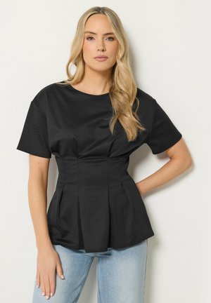 Lyshåret kvinde med løse bølger bærer en tætsiddende sort kortærmet top og lyseblå jeans, og står mod en ensfarvet hvid baggrund.
