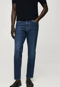 Jeans de mezclilla azul oscuro con corte recto, diseño de cinco bolsillos y costuras visibles. Combinados con sandalias negras tipo slip-on.