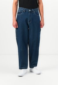 Vīrietis valkā brīvus tumši zilos džinsus ar sānu kabatām un baltiem sporta apaviem, stāvot pret baltu fona.