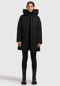 Melnā polsterētā jaka ar kapuci, kas ir ar kažokādas oderi, priekšējo rāvējslēdzēju un sānu kabatām. Uzvilkta virs melnajām legingām un potīšu zābakiem.