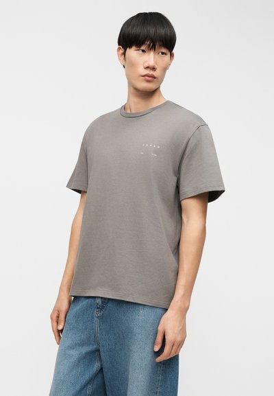 Γκρι βαμβακερό T-shirt με στρογγυλή λαιμόκοψη και κοντά μανίκια, με διακριτικό μπροστινό λογότυπο. Συνδυάζεται με φαρδιά μπλε τζιν παντελόνια.