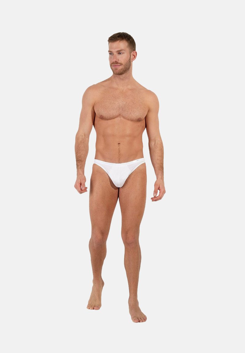 Slip bikini in cotone bianco, design a vita bassa, texture morbida, silhouette aderente, indossati da un modello maschile in piedi a piedi nudi su uno sfondo semplice.