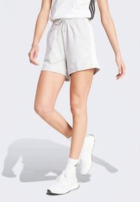 Άτομο με ανοιχτό γκρι σορτς Adidas με λευκές ρίγες, λευκές κάλτσες και λευκά αθλητικά παπούτσια, στέκεται με το ένα πόδι σταυρωμένο.