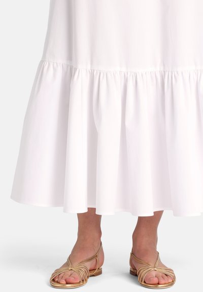 Den nederste del af en hvid lagdelt kjole båret med gyldne flade sandaler med remme på bare fødder mod en hvid baggrund.