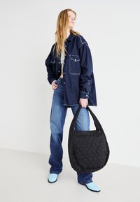 Κουβερτισμένη μαύρη τσάντα με στρογγυλό σχήμα, διαθέτοντας μαλακή υφή και μία μόνο χειρολαβή. Φορεμένη με ένα τζιν μπουφάν και μπλε τζιν.