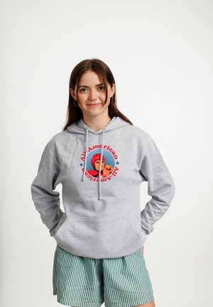 Junge Frau trägt einen grauen Hoodie mit dem Text „All-American“ und einer Boxer-Grafik, kombiniert mit grünen gestreiften Shorts, lächelt und hat die Hände in den Taschen.