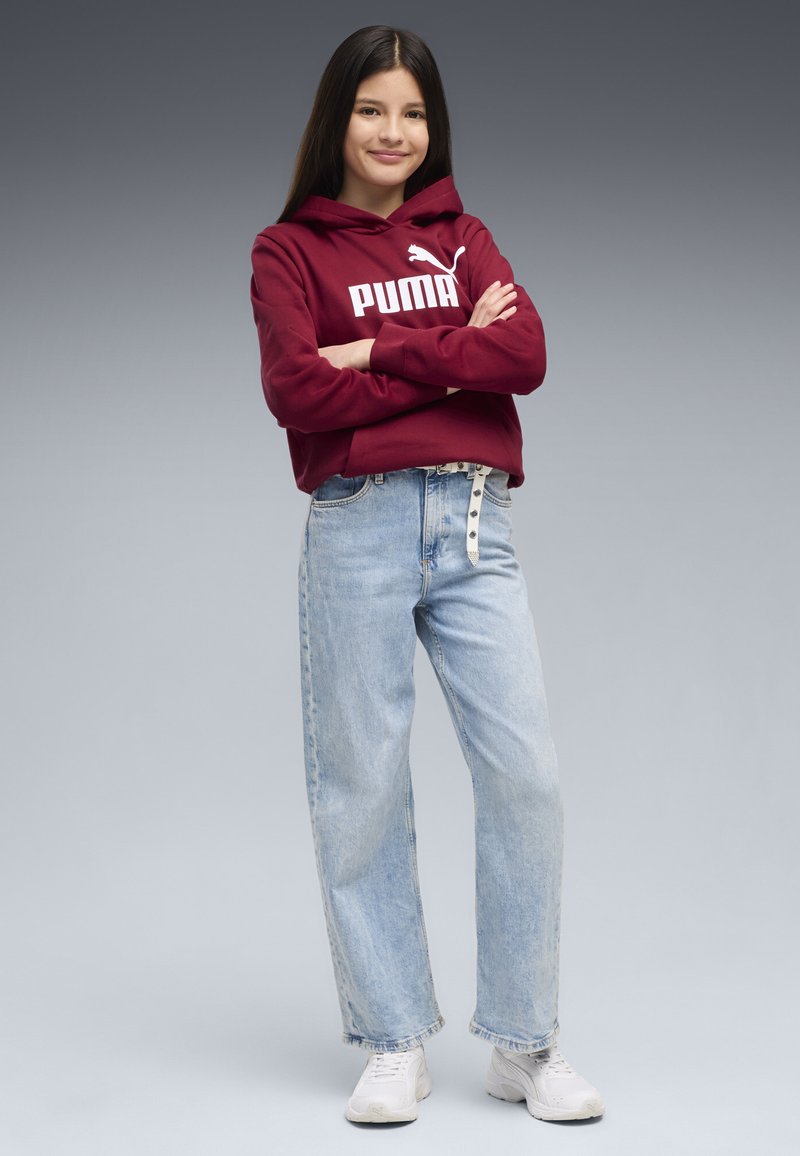 Jeune fille aux longs cheveux foncés, debout les bras croisés, portant un sweat à capuche Puma rouge, un jean bleu clair et des baskets blanches, sur fond gris.