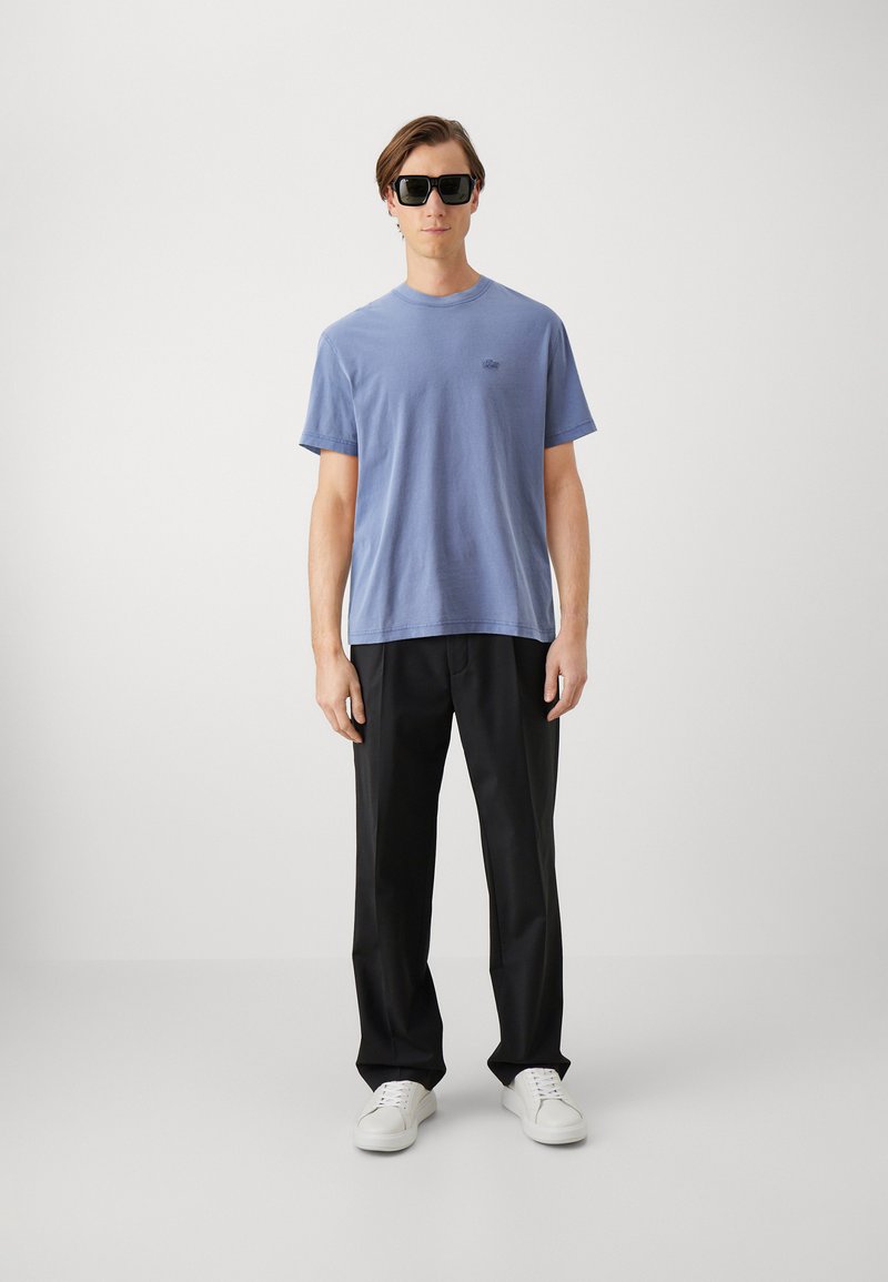 Camiseta de algodón azul de cuello redondo, pantalones negros y zapatillas blancas. Los detalles notables incluyen un pequeño logo en la camiseta y gafas de sol oscuras.