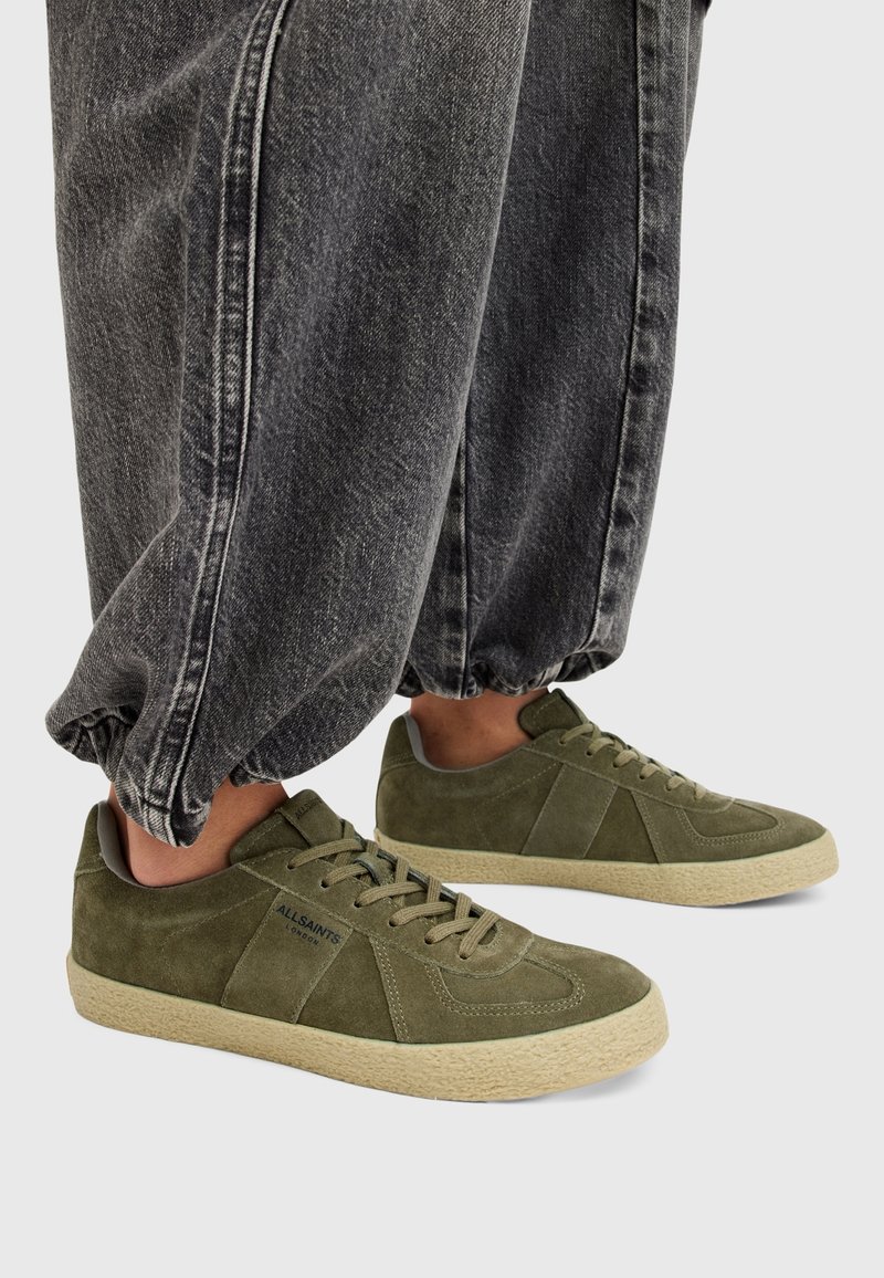 Baskets en suède vertes avec semelles beige, portées avec un pantalon en jean noir ample resserré aux chevilles sur fond clair.