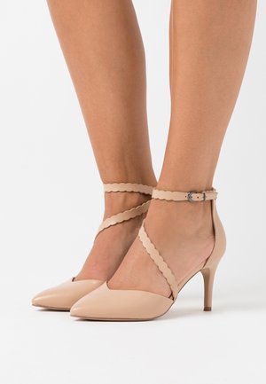 Chaussures à talons beiges avec des brides découpées à la cheville portées aux pieds, présentées sur un fond blanc uni.