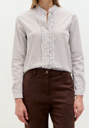 Mujer con camisa blanca abotonada con rayas verticales marrones y volantes, metida en pantalones de tiro alto de color marrón oscuro.