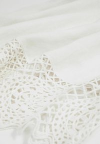 Witte stof met ingewikkelde kantafwerking met geometrische en cirkelvormige patronen, gevouwen getoond op een vlakke ondergrond.