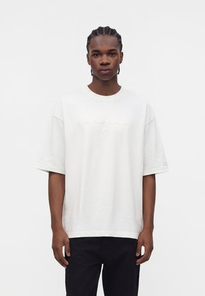 Jeune homme aux cheveux tressés, portant un t-shirt Jordan blanc oversize et un pantalon noir, debout devant un fond blanc uni.