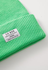 Zöld kötött sapka bordás textúrával. Fehér címkével rendelkezik, rajta a "LIL' BOO" felirattal és a márkainformációval. Puha, rugalmas anyag, hideg időjáráshoz alkalmas.