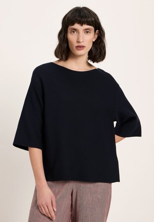 Frau mit kurzen dunklen Haaren trägt ein locker sitzendes schwarzes Oberteil mit Ellbogen-langen Ärmeln und eine hellrosa Hose mit weitem Bein, steht vor einem schlichten Hintergrund.