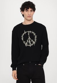 Black ribbed sweater featuring a peace symbol outlined in barbed wire. Crew neck and long sleeves, paired with black pants.
