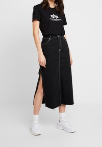 Falda de mezclilla negra con una gran abertura, dobladillo desgastado y detalles de costura blanca. Combinada con una camiseta gráfica negra y zapatillas blancas.